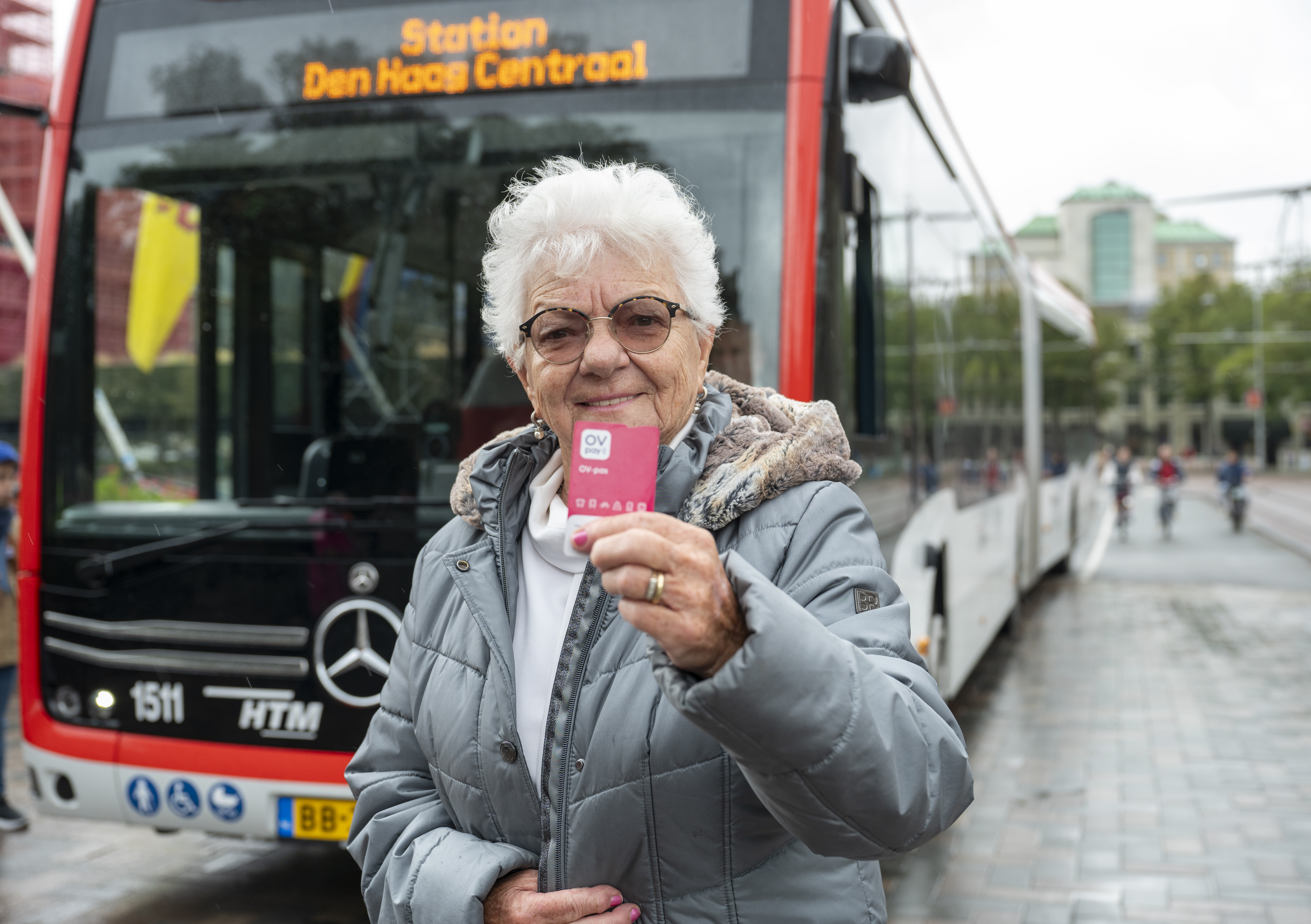Oude vrouw met bril staat voor een HTM bus met een nieuwe roze OV-pas in haar hand