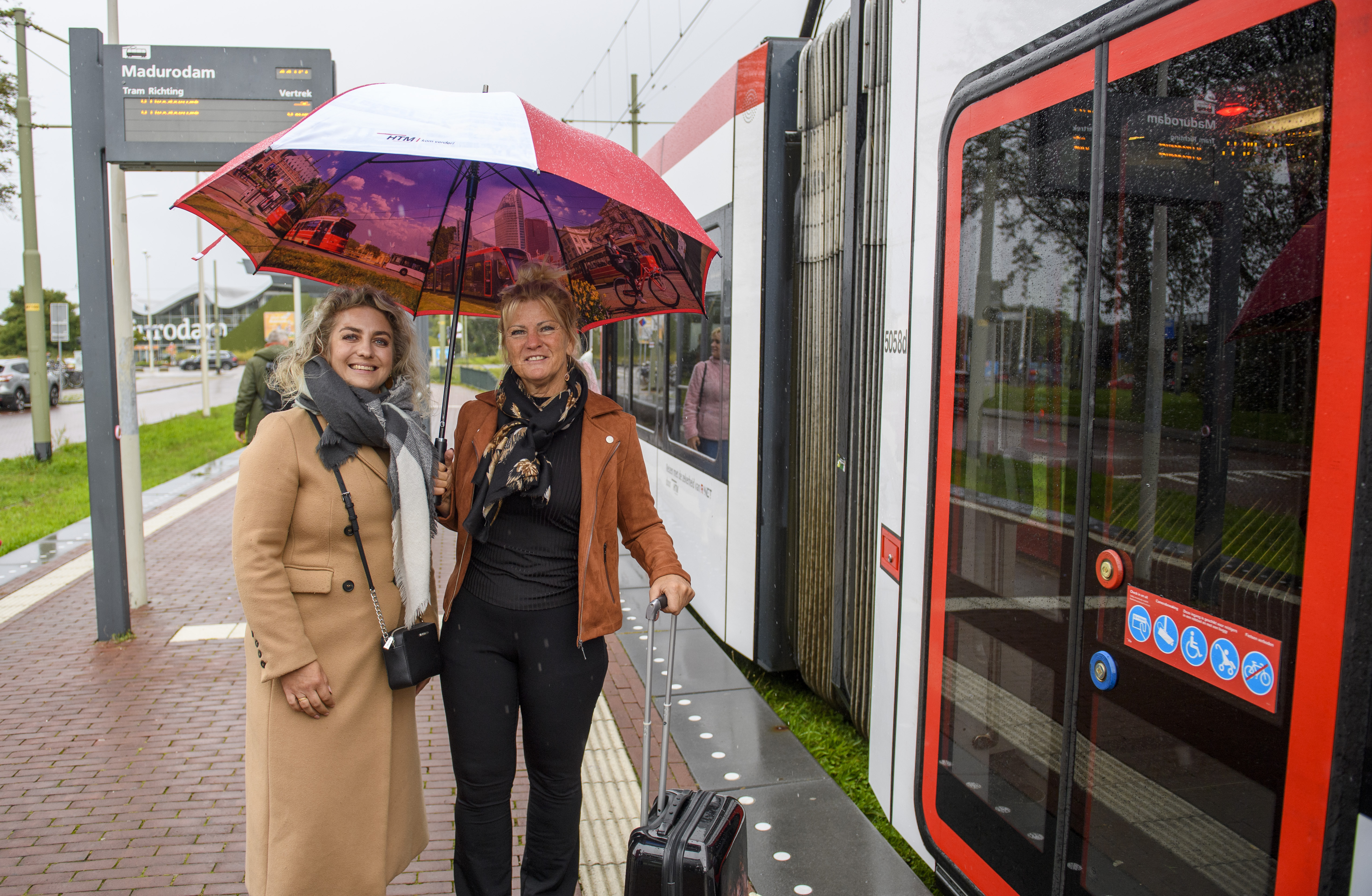 Reizigers staan op de halte onder een paraplu naast een HTM tram en lachen naar de camera