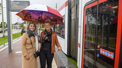Reizigers op halte naast tram Reizigers staan op de halte onder een paraplu naast een HTM tram en lachen naar de camera